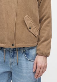 Main droite reposant sur le côté portant une veste en velours côtelé marron avec une poche à rabat à bouton-pression et un jean en denim bleu clair.
