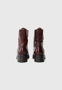 Botas de cuero marrón hasta el tobillo con un acabado suave, punteras redondas y un tacón grueso negro; incluye lengüetas para facilitar su puesta.