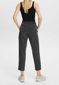 Femme vue de dos portant un débardeur noir, un pantalon cropped gris foncé avec taille élastique, des baskets blanches, debout les mains dans les poches.