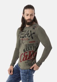 Olivegrünes Langarm-T-Shirt mit auffälligen schwarzen und roten Grafiken, darunter Text. Hat einen runden Halsausschnitt und eine schmal geschnittene Passform.