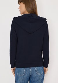 Donna di spalle che indossa una felpa con cappuccio blu navy e jeans blu su uno sfondo light semplice.