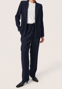 Námořnický blazer s přiléhavým střihem, s jedním knoflíkem a kapsami, doplněný o kalhoty s elastickým pasem ve stejné barvě. Černé špi�čaté boty.
