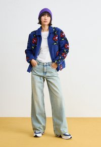 Blue, fleece jacket with floral embroidery; white t-shirt; light wash, wide-leg jeans; purple beanie; sporty shoes with layered accents.