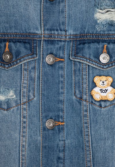 Chaqueta de mezclilla en azul claro con botones plateados, detalles desgastados, costuras naranjas y un parche bordado de un oso de peluche etiquetado como "TOY".