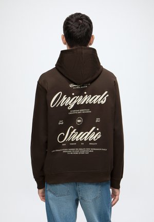 Man draagt een donkerbruine hoodie met witte tekst ""Originals Studio"" en extra kleinere tekst op de achterkant, gecombineerd met blauwe spijkerbroek.