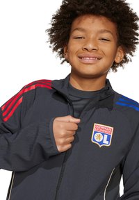 Donkergrijze jas met rode en blauwe schouderstrepen. Voorzien van een rits aan de voorkant en een geborduurd teamlogo. Zachte stoftextuur.