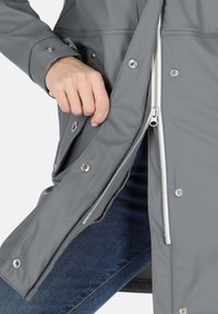 Giacca impermeabile grigia con una cerniera visibile, dettagli con bottoni a pressione e una mano nella tasca. Texture liscia e design su misura.