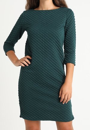 Femme portant une robe texturée vert foncé longueur genou avec des manches trois-quarts, une main sur la hanche et l'autre détendue le long du corps.
