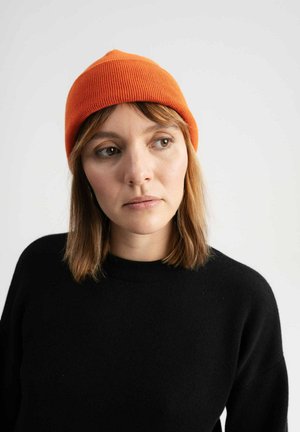 Tangerkleurige geribbelde breimuts met een taps toelopende top, gecombineerd met een zwarte gestructureerde trui, die een eenvoudig en schoon design heeft tegen een neutrale achtergrond.