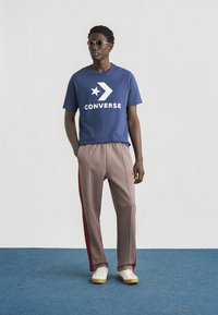 Μπλε ναυτικό βαμβακερό μπλουζάκι με μεγάλο λευκό λογότυπο "CONVERSE" και αστέρι, συνδυασμένο με καρό παντελόνια με κόκκινες λεπτομέρειες. Λευκά αθλητικά παπούτσια ολοκληρώνουν το σύνολο.