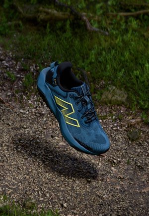 Chaussure de trail running New Balance bleue et jaune suspendue au-dessus d'un chemin de gravier avec de l'herbe verte en arrière-plan.