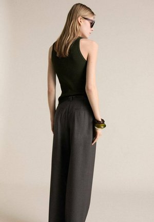 Pantaloni neri a gamba larga con una finitura testurizzata e una canotta aderente a coste di un verde scuro. Gli accessori includono braccialetti impilati su un polso.