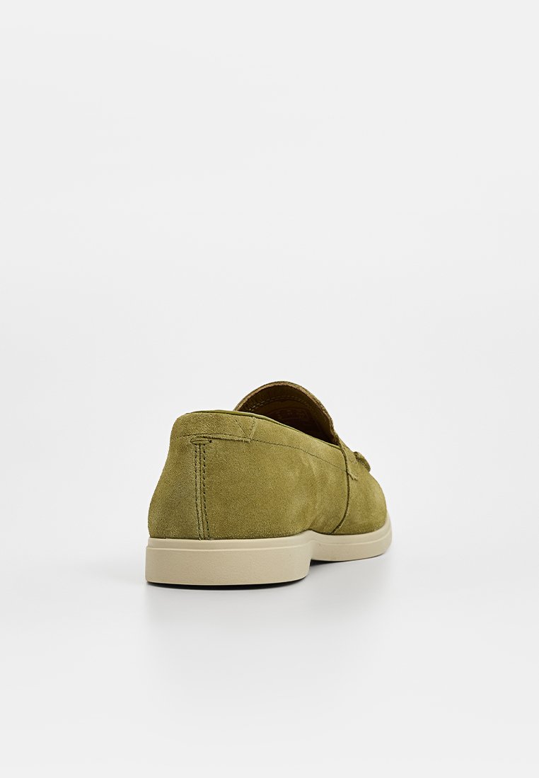 Des chaussures à enfiler en suede vert avec une semelle plate beige, présentant une texture lisse et un détail de couture minimal au niveau du talon.