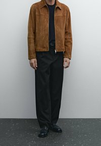 Brown suede jacket with a plain design, stand collar, and zipper closure, paired with black trousers and black shoes on a textured surface.