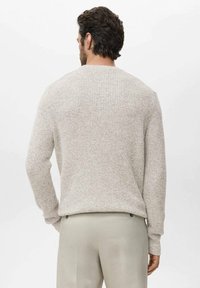 Man met donker haar die een lichtbeige gestructureerde gebreide trui en lichtgrijze broek draagt, staand met de rug naar de camera.