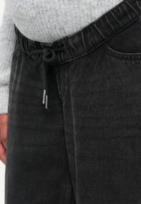 Črne denim hlače z elastiko v pasu in vrvico. Tekstura je gladka, z diskretnimi variacijami v zaključku tkanine.