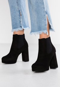 Botines de ante negro con un diseño de tacón grueso, que cuentan con paneles laterales elásticos y una plataforma negra brillante. Parte inferior de mezclilla clara deshilachada.