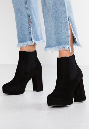Botines de ante negro con un diseño de tacón grueso, que cuentan con paneles laterales elásticos y una plataforma negra brillante. Parte inferior de mezclilla clara deshilachada.