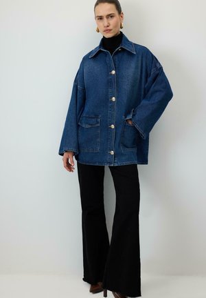 Kobieta w dużej, niebieskiej kurtce jeansowej z dużymi kieszeniami, czarnym golfem, czarnych rozkloszowanych spodniach i brązowych butach na obcasie, stoi przy białej ścianie.