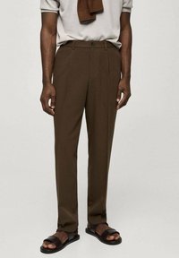 Braune, maßgeschneiderte Hose aus glattem Stoff mit flacher Vorderseite, Seitentaschen und Knopfverschluss. Kombiniert mit einem hellen Oberteil und Sandalen.