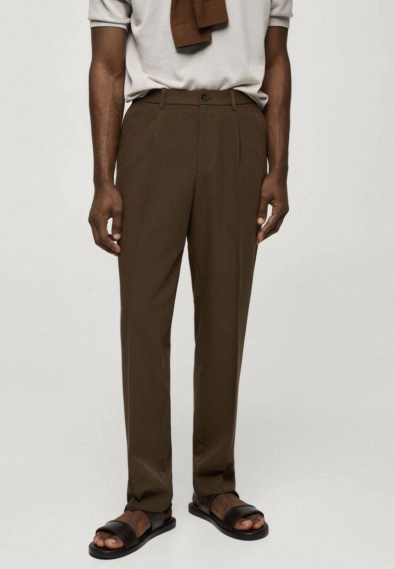 Braune, maßgeschneiderte Hose aus glattem Stoff mit flacher Vorderseite, Seitentaschen und Knopfverschluss. Kombiniert mit einem hellen Oberteil und Sandalen.