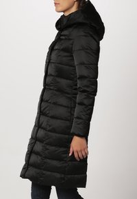 Schwarze Steppjacke mit Kapuze, ausgestattet mit einem gesteppten Design und vertikalen Nähten. Lange Länge, glatte Textur und ein zwei-Wege-Reißverschluss.