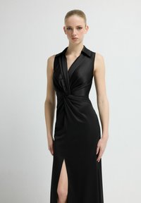 Czarna sukienka bez rękawów z gładkiej tkaniny, z kopertowym krojem z marszczeniem w talii i rozcięciem do uda po jednej stronie.