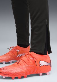 Chaussures de football Puma orange vif portées avec un pantalon noir muni de fermetures éclair latérales, présentées sur un fond neutre.