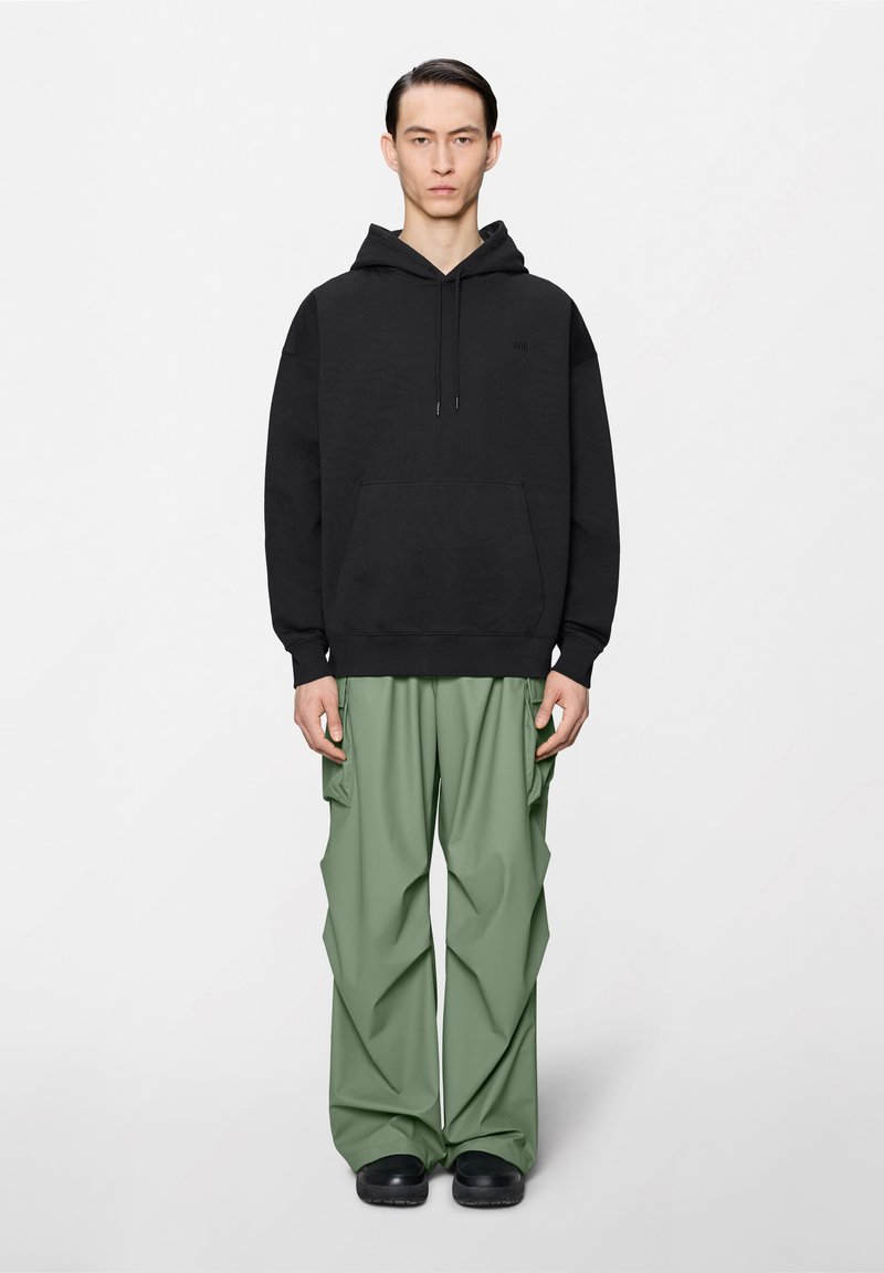 Homme debout portant un sweat à capuche noir, un pantalon cargo ample vert clair et des chaussures noires sur un fond blanc uni.