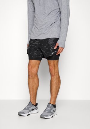 Mann steht und trägt ein graues Langarmshirt, schwarze sportliche Shorts mit Muster und Nike-Logo sowie graue Nike-Laufschuhe vor weißem Hintergrund.