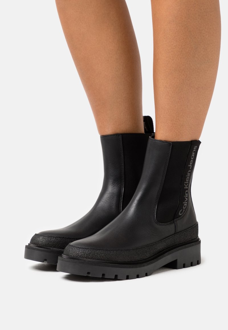 Bottines noires à semelles épaisses portées sur jambes nues, avec des empiècements en élastique sur les côtés et la marque "Calvin Klein Jeans" sur le côté.