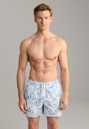 Shirtless man with short light brown hair wearing white and blue patterned swim shorts standing against a plain gray background.