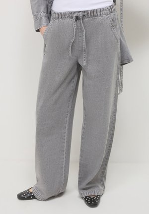 Persona che indossa pantaloni in denim grigio a vita alta e larghi con cordoncino, abbinati a ballerine nere con borchie e a una giacca di denim grigia coordinata.