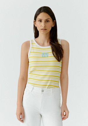 Femme aux longs cheveux foncés portant un haut sans manches rayé jaune et blanc avec un texte bleu et un pantalon blanc taille haute, debout sur un fond clair.