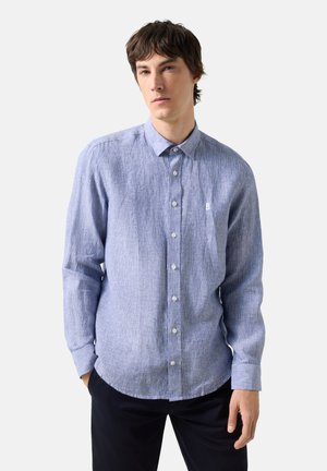 Junger Mann, der ein hellblaues Hemd mit langen Ärmeln und feiner Textur trägt, steht vor einem einfachen weißen Hintergrund.