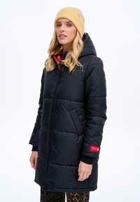 Čierna prešívaná bunda s kapucňou, so zipsom vpredu, bočnými vreckami a červeným logom ako akcentom. Skombinovaná so žltou čiapkou a patterned nohavicami.