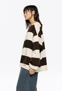 Mlada ženska z dolgimi temnimi lasmi, oblečena v širok rjav-bež pulover s črtami in ohlapne modre hlače, stoji vstran.