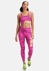 Rosa mönstrad sportbustier med gula accenter och logotyp, ihop med matchande rosa leggings som har en djärv logotyp. Modellen har på sig neonsneakers.