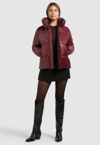 Bordeauxfarbene Steppjacke mit quiltierter Optik, Reißverschluss und abnehmbarer Kapuze. Getragen über einem schwarzen Minirock und schwarzen kniehohen Stiefeln.