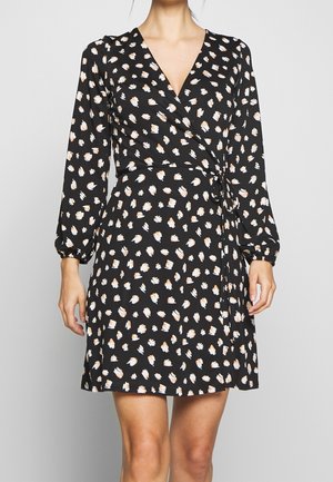 Sort slå-om-kjole med hvide og lysebrune abstrakte pletter, lange ærmer og et bundet taljebælte, båret af en stående person.