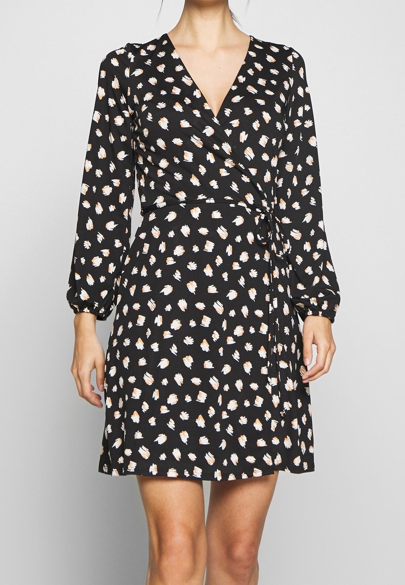 Robe portefeuille noire avec des taches abstraites blanches et marron clair, manches longues et ceinture nouée à la taille, portée par une personne debout.