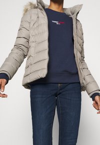 Person wearing a beige puffer jacket with fur hood over a navy Tommy Jeans sweatshirt and dark blue jeans.