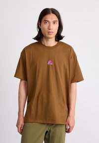Ung mann med skulderlangt svart hår iført en brun T-skjorte med en liten lilla ACG-logo og olivengrønne bukser, som står mot hvit bakgrunn.