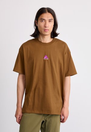 Young man with shoulder-length black hair wearing a brown T-shirt with a small purple ACG logo and olive-green pants, standing against white background.
