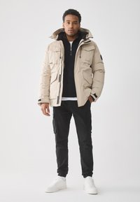 Uomo in piedi che indossa un giubbotto invernale beige con cappuccio, un maglione zip nero, pantaloni cargo neri e sneakers bianche su uno sfondo neutro.