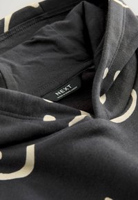 Sweatshirt noir avec un motif imprimé beige et une étiquette "NEXT Fabriqué au Bangladesh" visible à l'intérieur de l'encolure.
