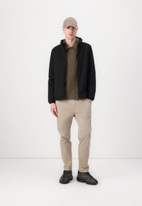 Sort zip-up jakke over en olivengrøn fleece polo, parret med beige bukser og sorte sneakers. Modellen bærer en tan hat med øre piercings.