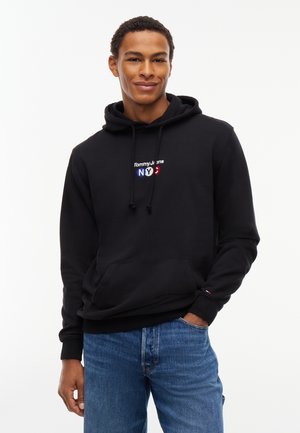 Junger Mann, der einen schwarzen Tommy Jeans Hoodie mit dem Text "NYC" und blaue Jeans trägt, lächelt mit einer Hand in der Tasche vor weißem Hintergrund.
