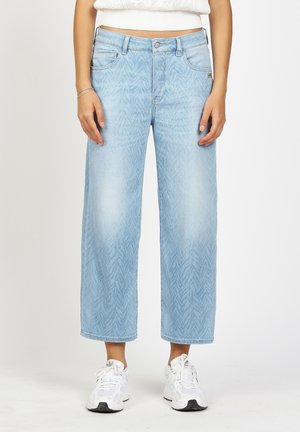 Person wearing light blue, wide-leg cropped jeans with subtle chevron pattern and white sneakers, standing against a plain background.