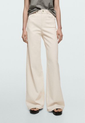 Personne portant un pantalon crème taille haute à jambes larges et des sandales plateformes noires, debout devant un fond clair uni.